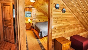 Una camera da letto mansardata accogliente, con soffitto spiovente, pareti in legno, un letto con coperta a quadri, un comodino con lampada e due cornici appese al muro.