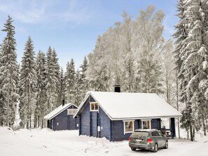 Une cabane dont le toit est recouvert de neige, entourée d’arbres chargés de neige, et une voiture garée devant.