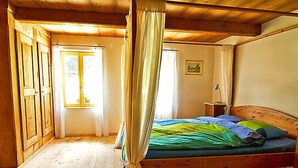 Una habitación con techo de madera, cama con colcha verde y azul, mesita de noche con lámpara y ventana con cortinas.