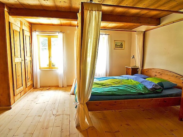 Une pièce avec un plafond en bois, un lit recouvert d’une couverture verte et bleue, une table de chevet avec une lampe et une fenêtre avec des rideaux.