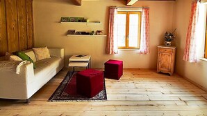 Una habitación acogedora con techo de madera, un sofá, una mesita, un reposapiés rojo y un ventanal con cortinas.