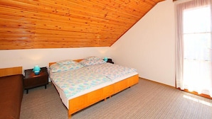 Un dormitorio con techo inclinado, una cama con ropa de cama floral, una mesita de noche y un ventanal con cortinas.