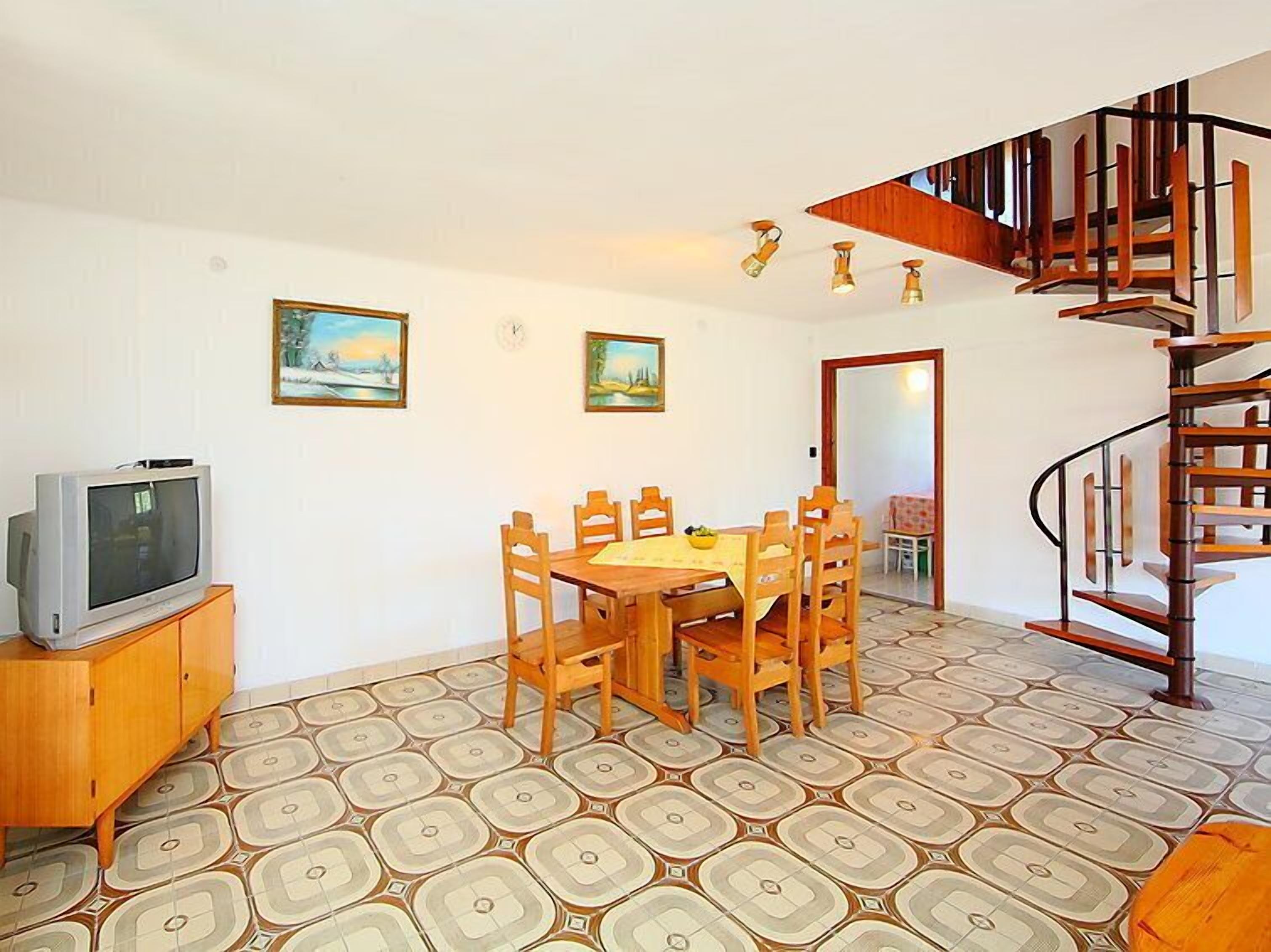 A dining area with a wooden table and chairs, a television on a cabinet, and a patterned floor.
