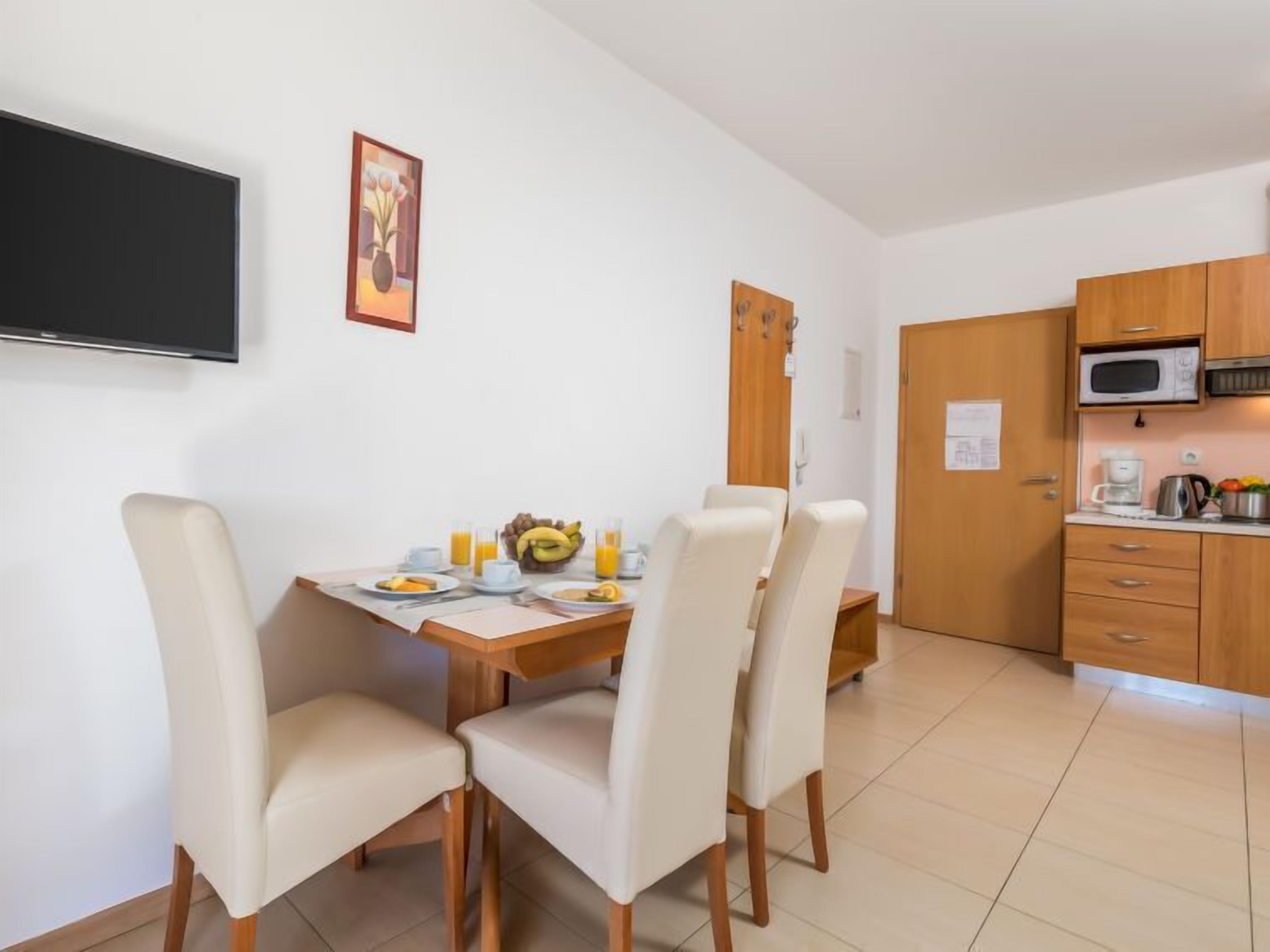 A dining area with a table set for two, chairs, a flat-screen TV mounted on the wall, and a kitchen area with a microwave and cabinets.
