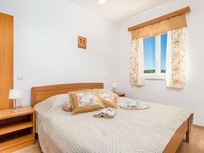 A bedroom with a wooden bed, a bedside table with a lamp, a window with floral curtains, and a small wall art piece.