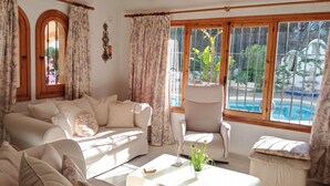 A spacious living room with a beige sofa, a white armchair, and a coffee table. A large window with a view of a swimming pool and greenery is visible. - Amadorio by Interhome (Jávea)