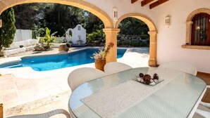 A pool area with a glass table and chairs, surrounded by arches and a stone wall. - Amadorio by Interhome (Jávea)