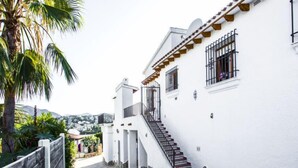 Un edificio blanco con una escalera de piedra que conduce a un balcón, rodeado de vegetación y vistas a un pueblo lejano.