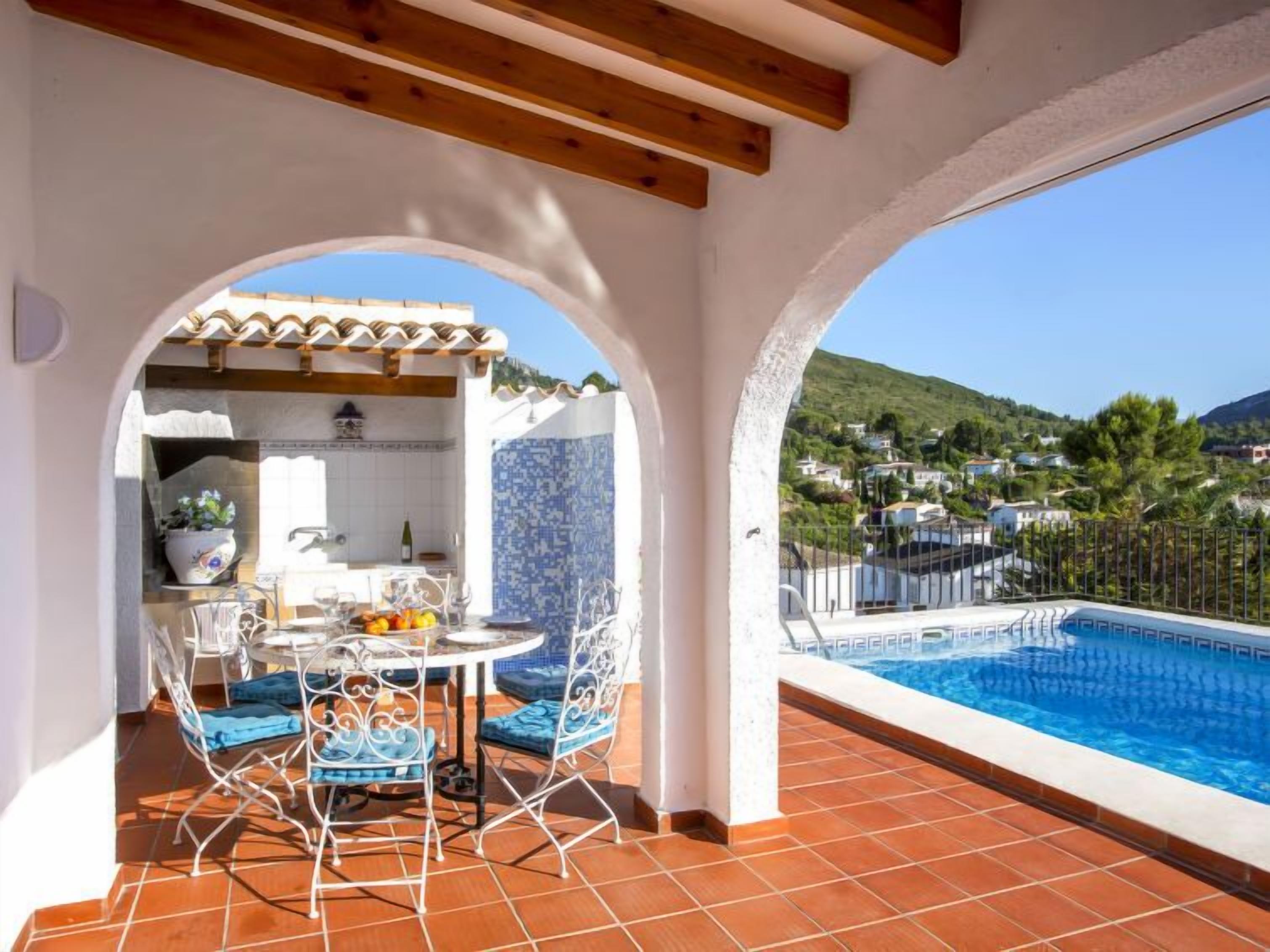A terrace with a pool, a dining area with a table and chairs, and a view of a hillside town.