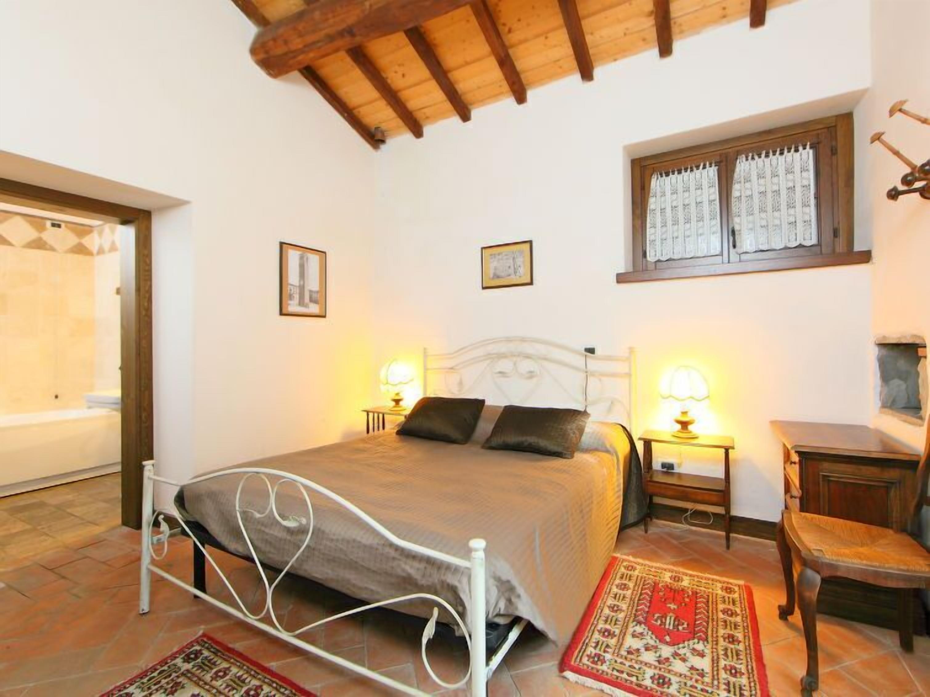 A bedroom with a bed, two bedside tables with lamps, a chair, a window with curtains, and a wooden ceiling.
