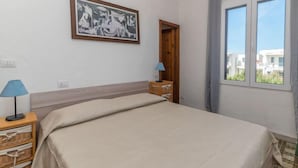 A bedroom with a bed, a nightstand, a lamp, a framed picture on the wall, and a window with a view of buildings. - Villa Selena by Interhome (Valledoria)
