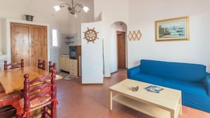 A living room with a blue sofa, wooden dining table, and chairs. There is a kitchen area with cabinets and a stove. A clock and a framed picture are on the wall. - Villa Selena by Interhome (Valledoria)