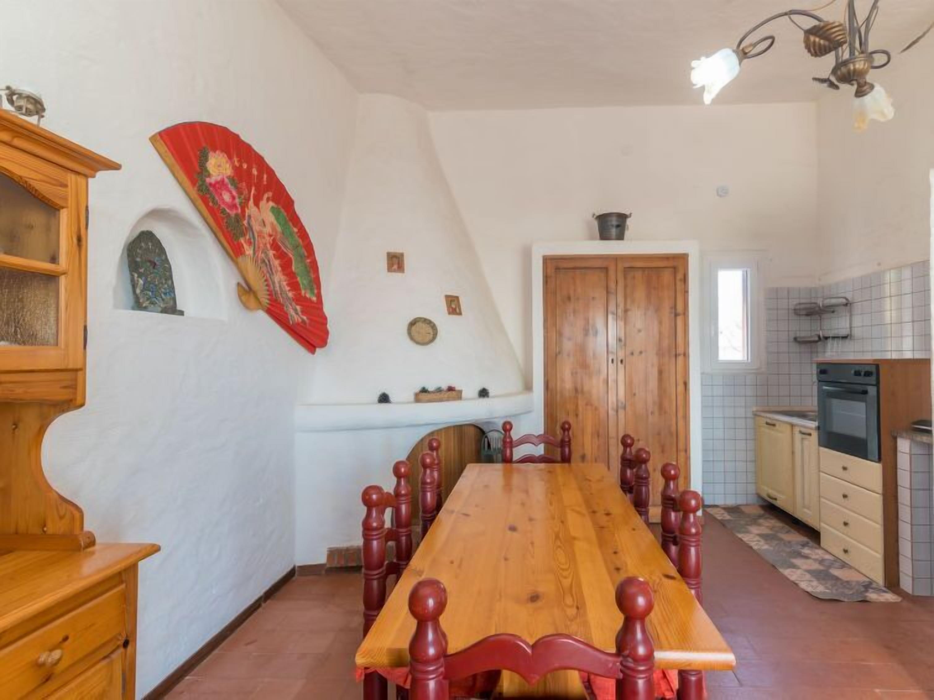 Een eetgedeelte met een houten tafel en stoelen, een keuken met witte tegels, en een rode decoratieve ventilator aan de muur.