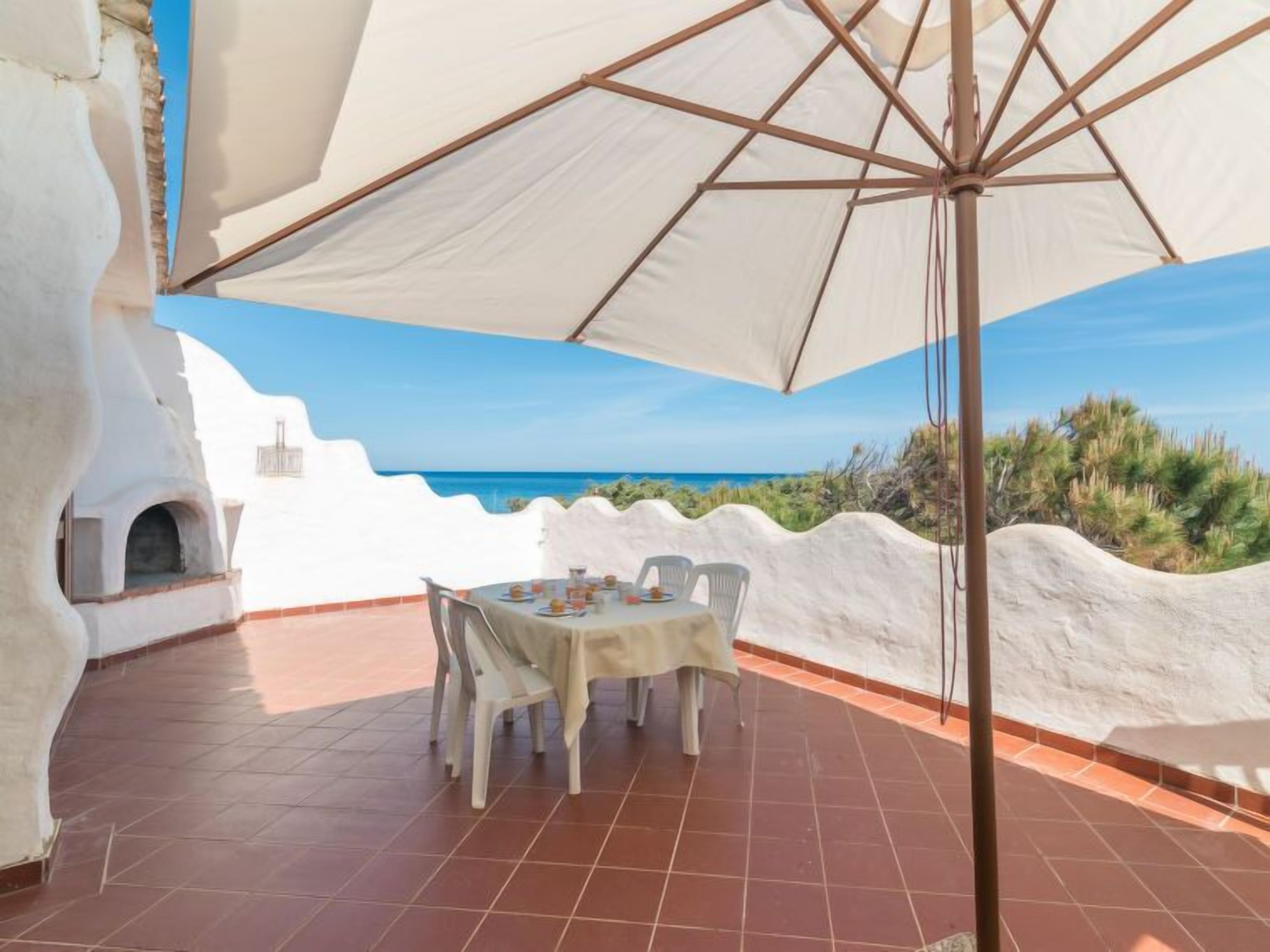 Een eetgedeelte met een tafel en stoelen onder een grote parasol, met uitzicht op de kust.