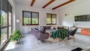 Un salon spacieux avec un plafond haut, de grandes fenêtres équipées de volets et un téléviseur moderne fixé au mur.