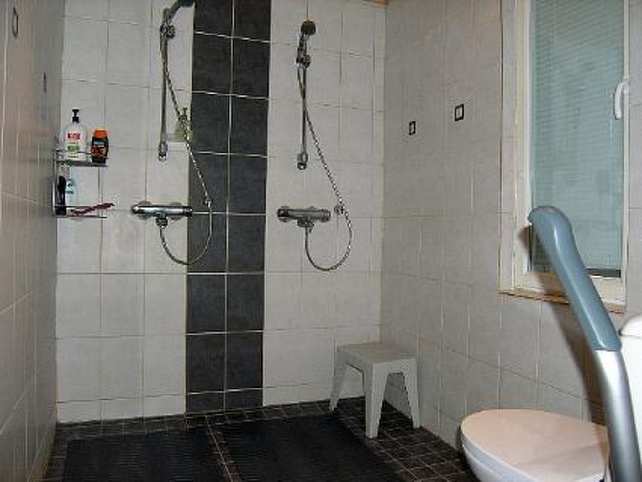 A bathroom with two showerheads, a toilet, a small stool, and a shelf with toiletries.