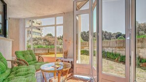 Una habitación iluminada por el sol con un ventanal de vidrio que da al exterior, mobiliario de mimbre y piso de baldosas.