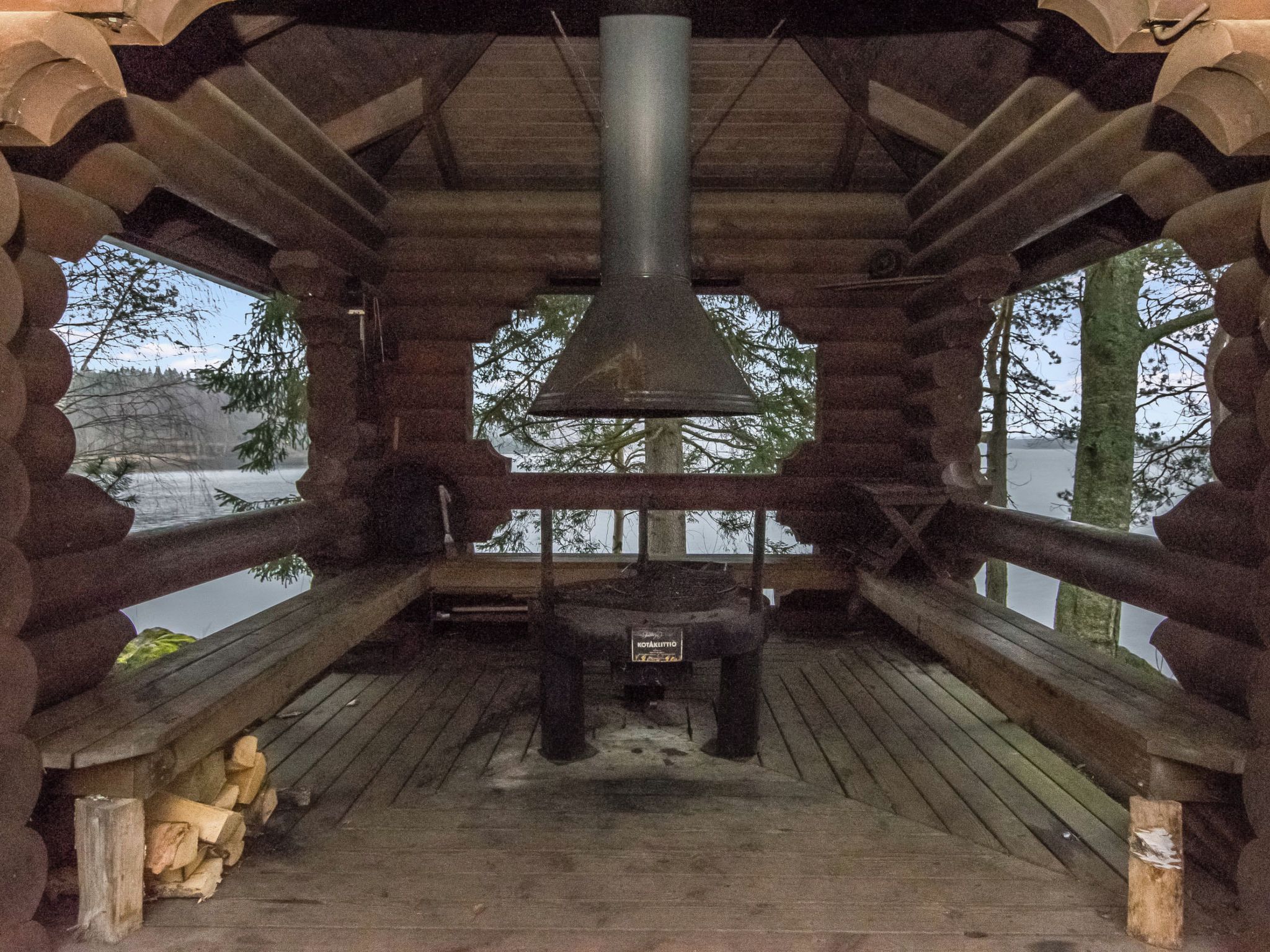 A wooden cabin with a central chimney, a wooden deck, and a view of a lake.