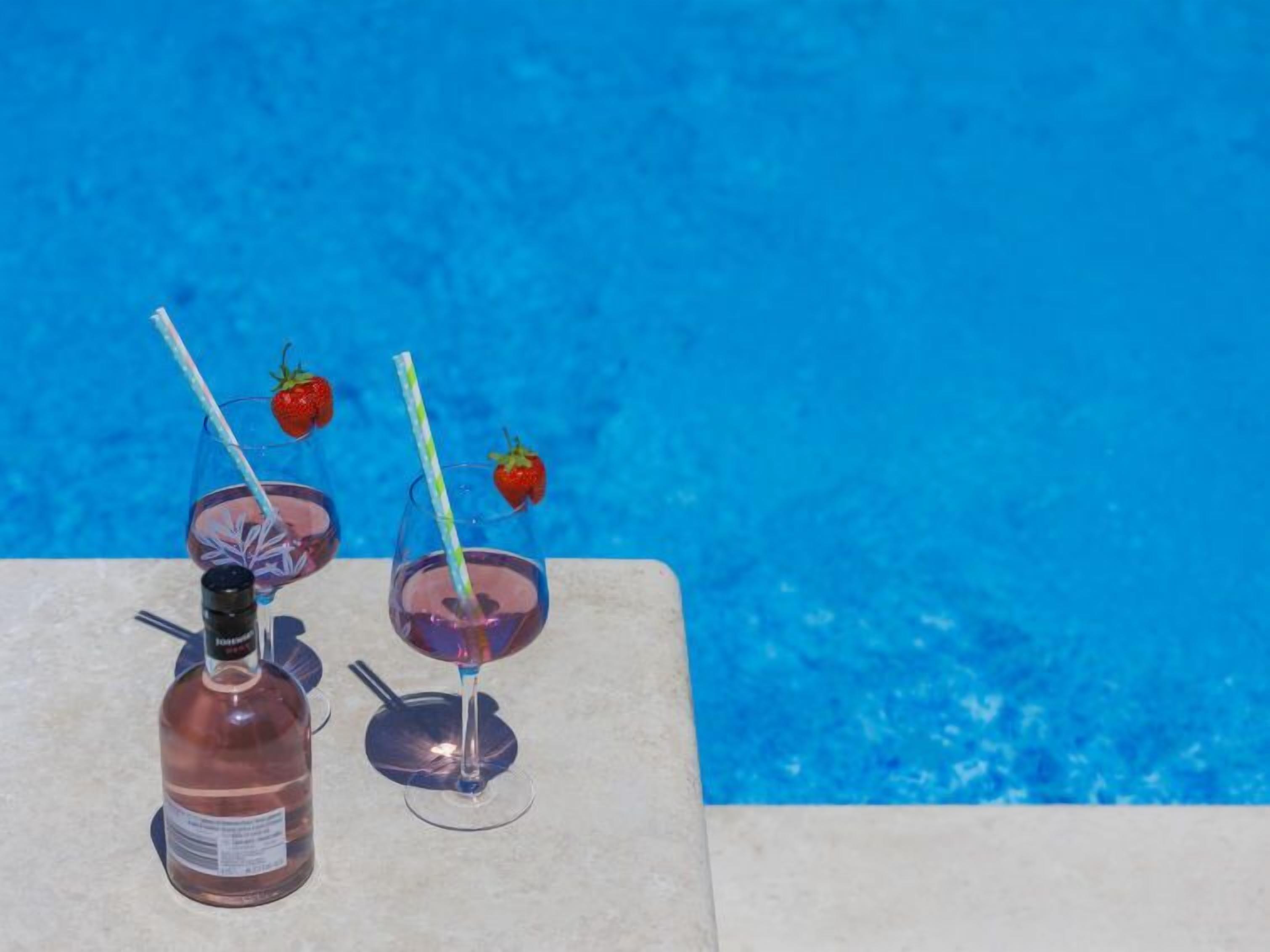 A poolside setting with a bottle of rosé wine and two glasses with straws and strawberries.