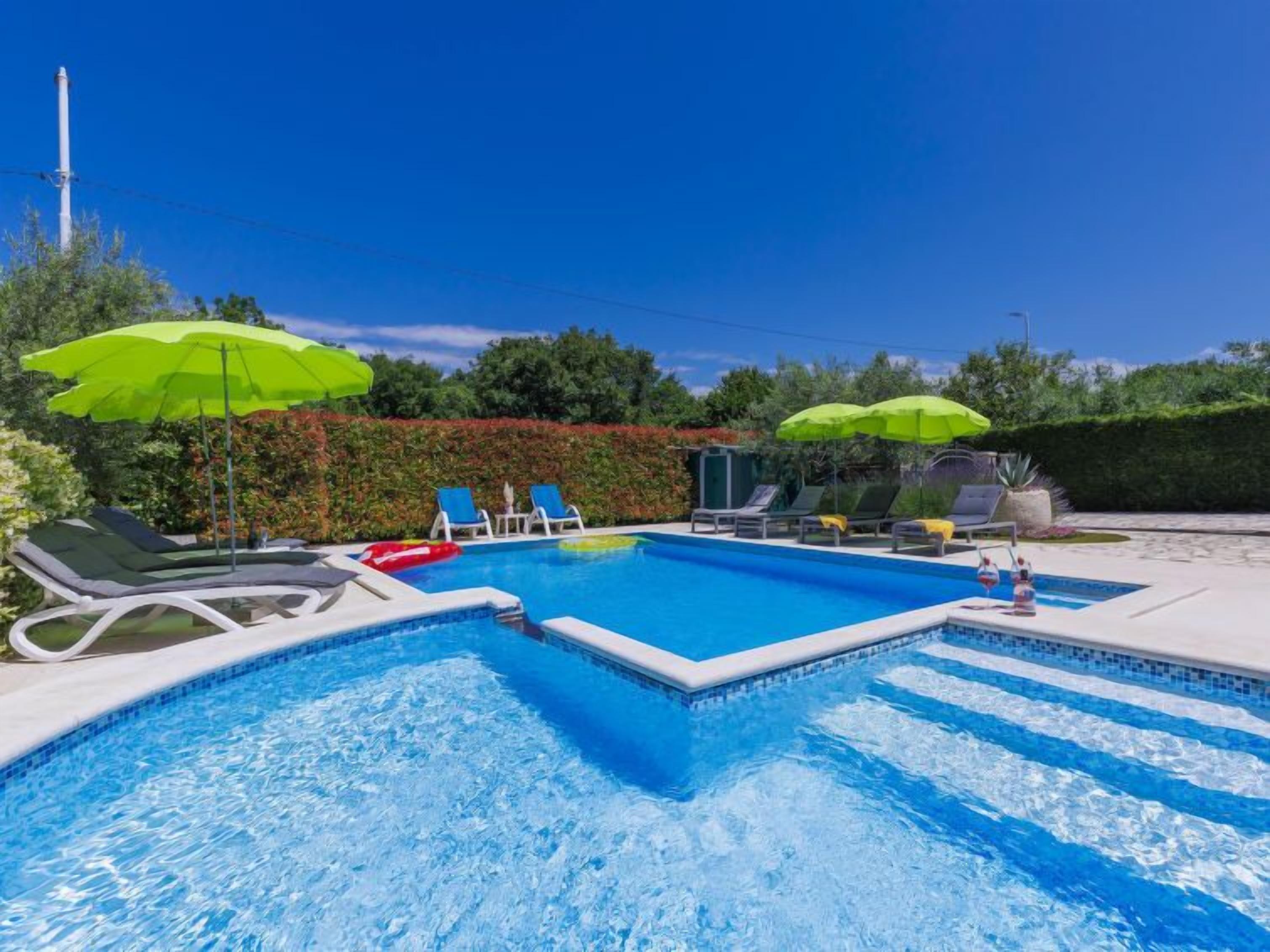 Ein Swimmingpool mit Liegestühlen und Sonnenschirmen, umgeben von viel Grün.