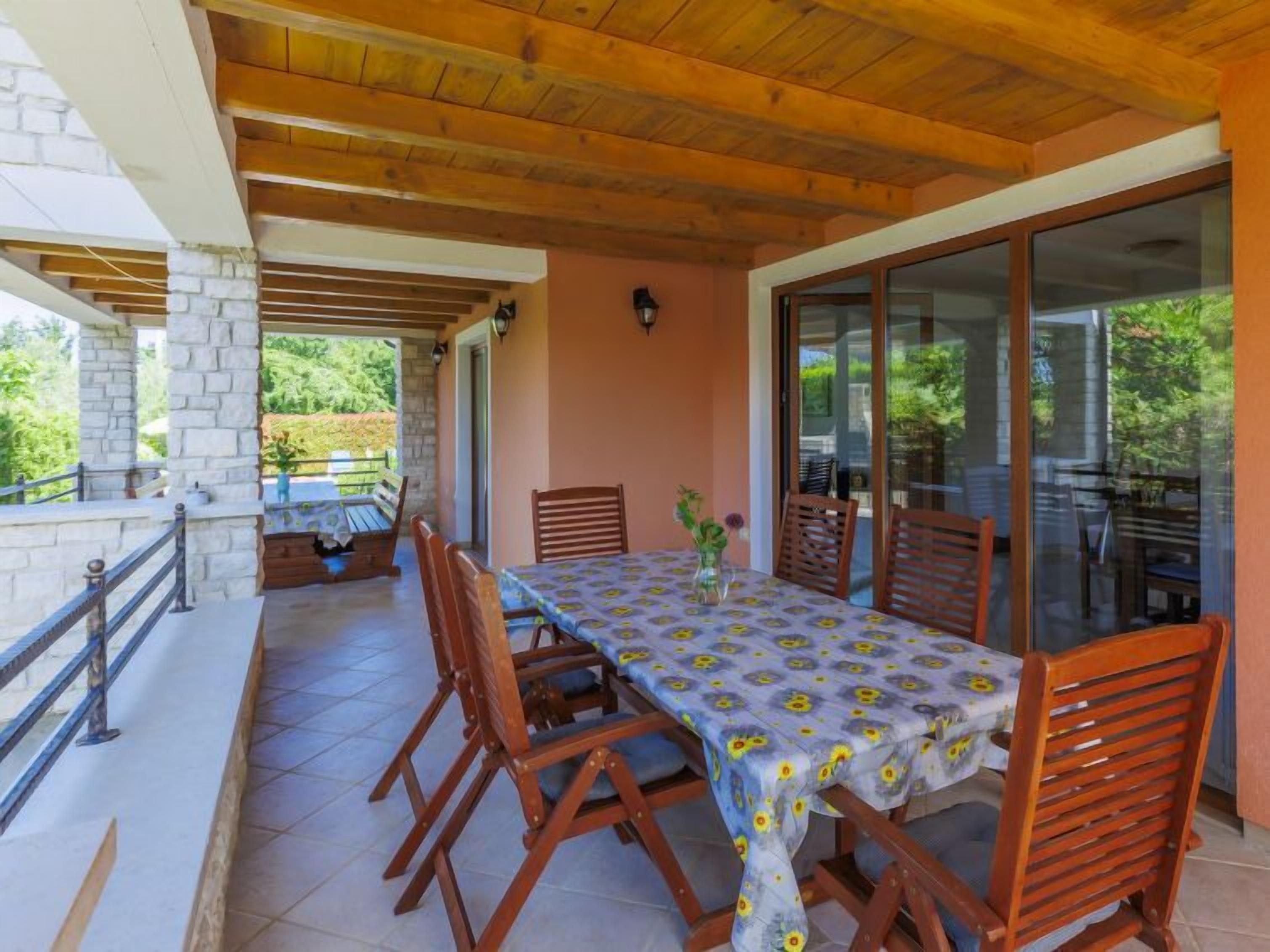 Une terrasse extérieure avec une table et des chaises, des piliers en pierre, des poutres en bois et une porte vitrée donnant sur un jardin.