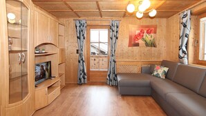 A cozy cabin interior with wooden walls and ceiling, a gray sofa, a glass cabinet with a TV, and curtains with a leaf pattern. - Katharina by Interhome (Aschau im Zillertal)