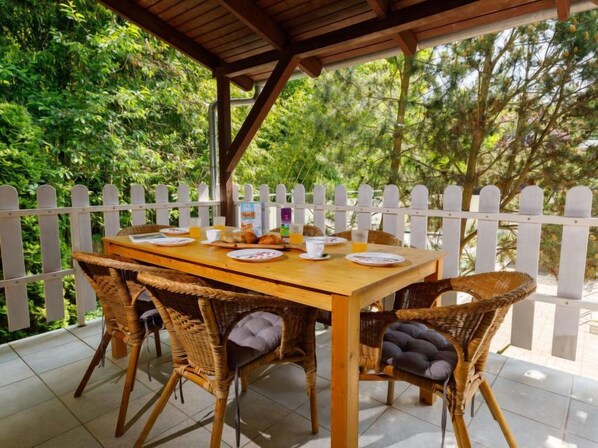 A wooden table set for breakfast with wicker chairs, a white picket fence, and lush greenery in the background.