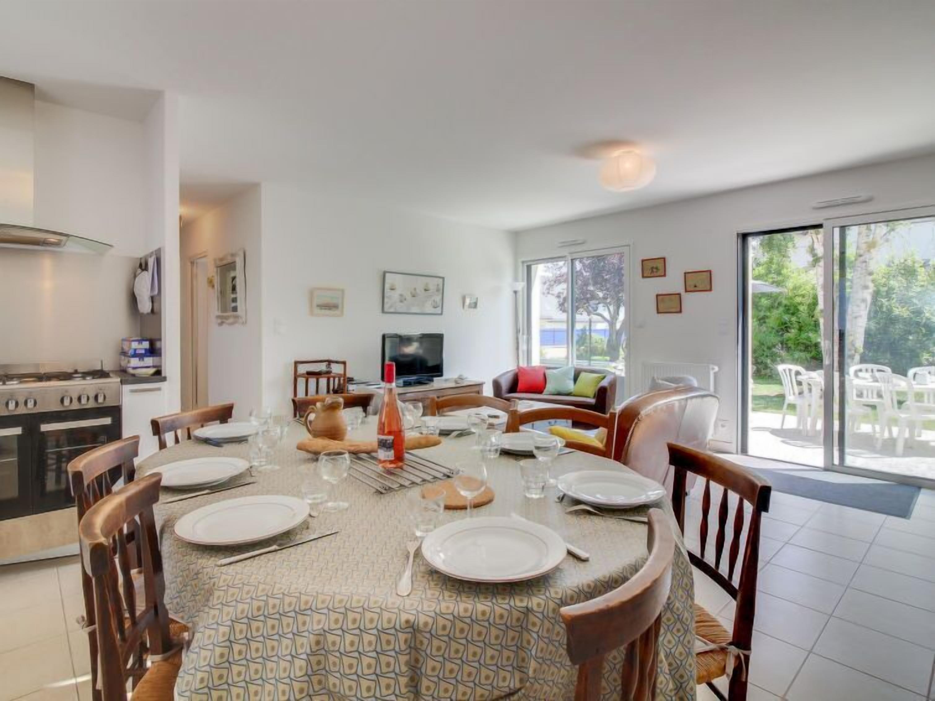Un coin repas avec une table ronde dressée pour six personnes, une cuisine équipée d’une cuisinière et d’un four, et un salon avec un téléviseur et un canapé.