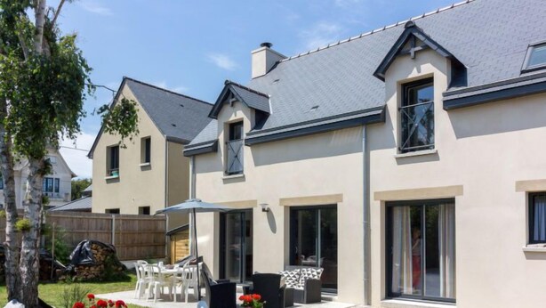 Ein modernes, zweistöckiges Haus mit Terrasse, überdachter Sitzgruppe und einem Garten mit roten Blumen.