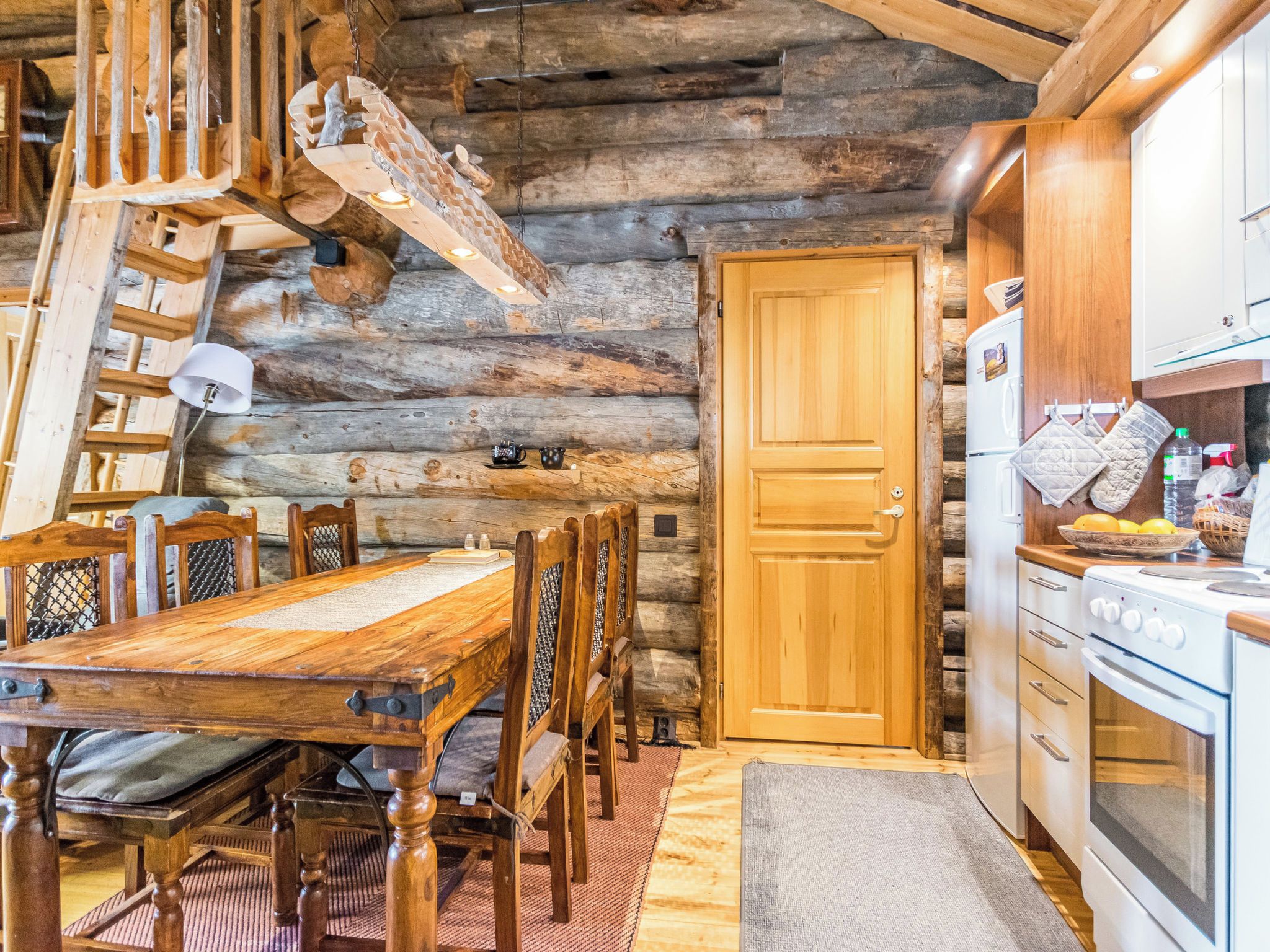 A rustic wooden interior with a dining table, chairs, a wooden ladder, a wooden door, and a kitchen area with a stove and cabinets.