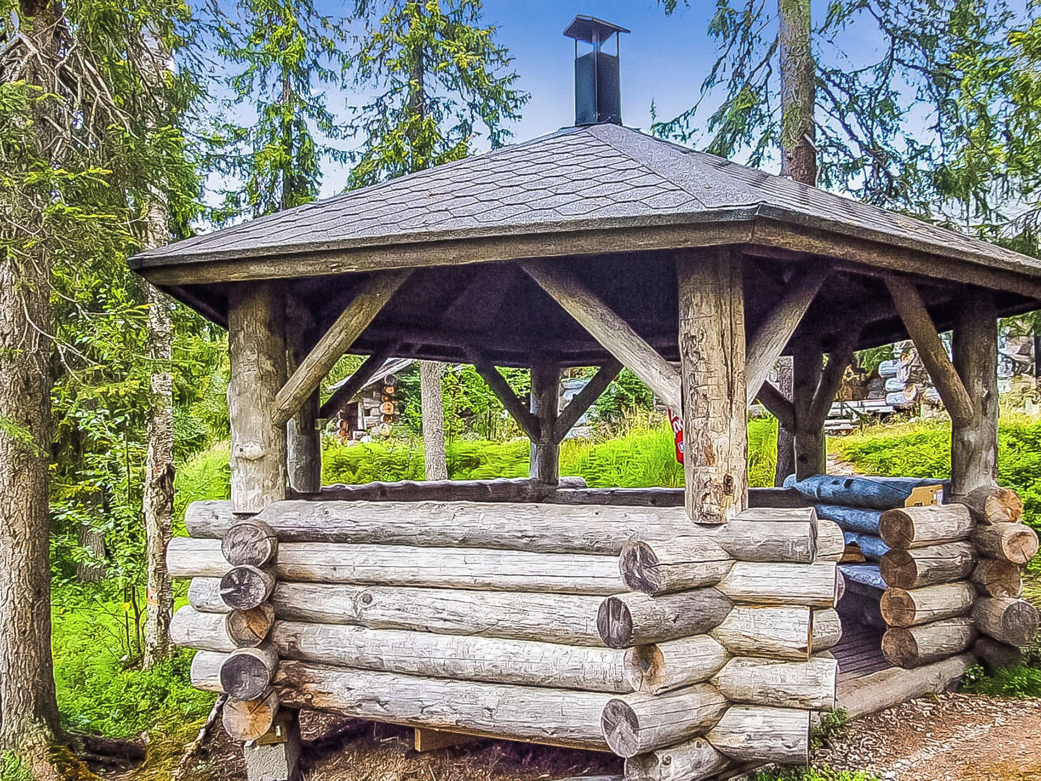 A rustic wooden gazebo with a sloped roof, surrounded by lush greenery and trees.