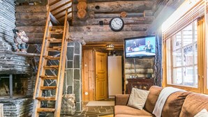 Tunnelmallinen hirsihuvilan sisustus, jossa on kivitakka, puinen tikapuu, sohva ja televisio.