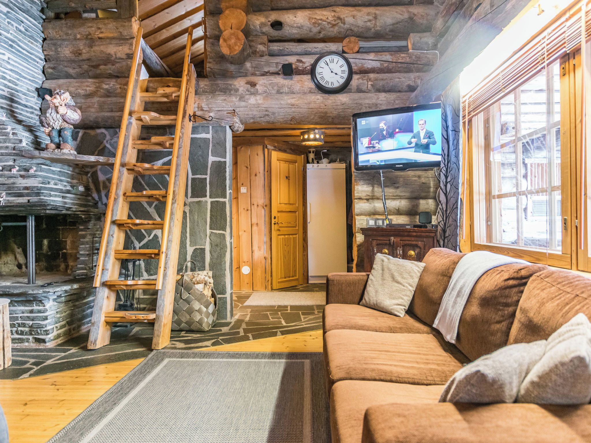 Interior acogedor de una cabaña de madera con chimenea de piedra, escalera de madera, sofá y televisor.