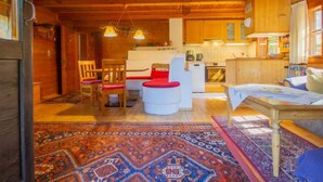 A cozy cabin interior with a wooden ceiling, a kitchen area, a dining space with a red ottoman, and a patterned rug.