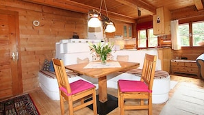 A wooden cabin interior with a dining table, chairs, a kitchen area, and a window.