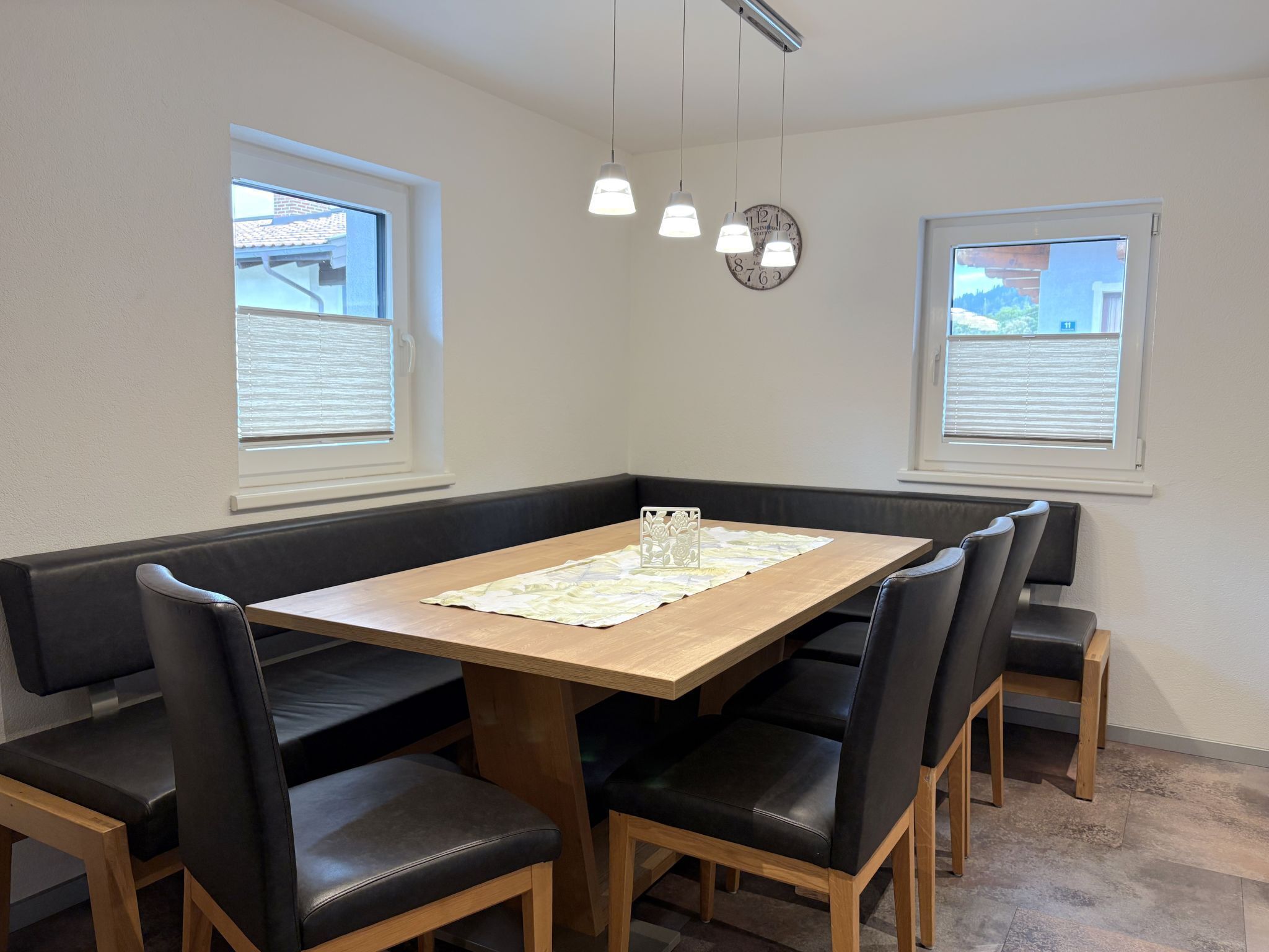 A dining area with a wooden table, black cushioned chairs, pendant lights, and windows with blinds.