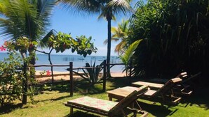 Un patio frente a la playa con sillones reclinables de madera y una mesa, rodeado de plantas tropicales y una valla de madera.