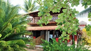 Un edificio de madera de dos pisos con balcón, rodeado de abundante vegetación y palmeras.
