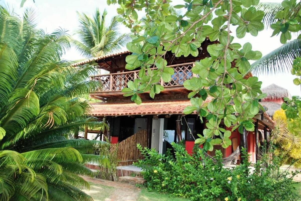 A two-story wooden building with a balcony, surrounded by lush greenery and palm trees.