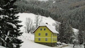 A yellow building with a sloped roof, surrounded by snow-covered trees and a mountainous backdrop.