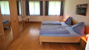 A room with a wooden floor, two beds with checkered bedding, a wooden desk, a mirror, and a window with curtains.