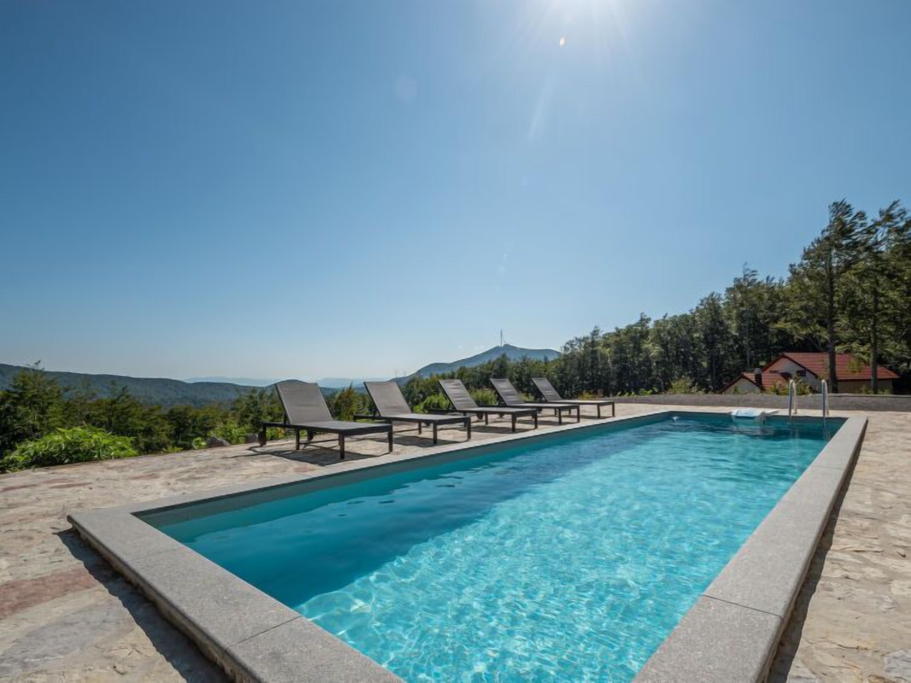 A rectangular swimming pool with clear blue water, surrounded by a stone deck. There are six lounge chairs aligned along the pool's edge. The pool is set against a backdrop of green trees and distant mountains under a clear blue sky.