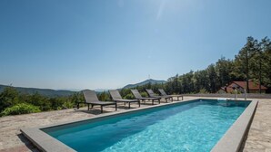 Ein rechteckiger Swimmingpool mit klarem, blauem Wasser, umgeben von einer Steindeck. Sechs Liegestühle sind entlang des Poolrands aufgereiht. Im Hintergrund erstrecken sich grüne Bäume und ferne Berge unter blauem Himmel.