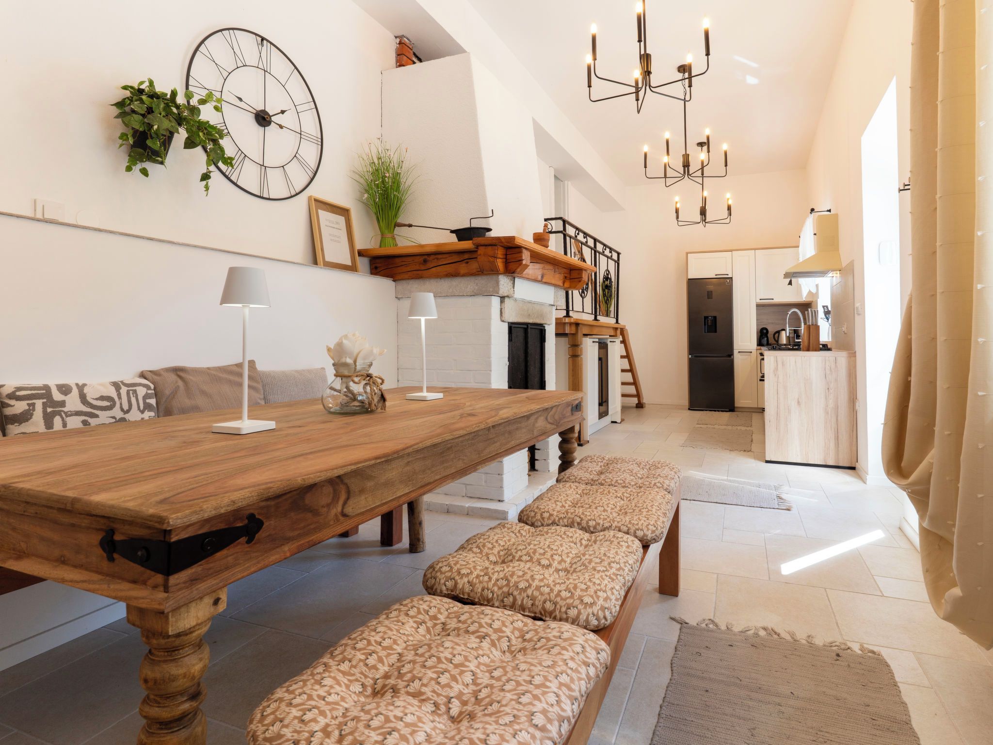 A spacious living area with a wooden dining table, benches, a large wall clock, and a modern kitchen in the background.