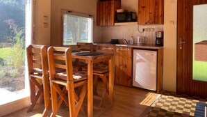 Een compacte keuken met houten kastjes, een magnetron, koffiezetapparaat en een kleine koelkast. Er is een eettafel met stoelen en een raam dat zorgt voor natuurlijk licht.