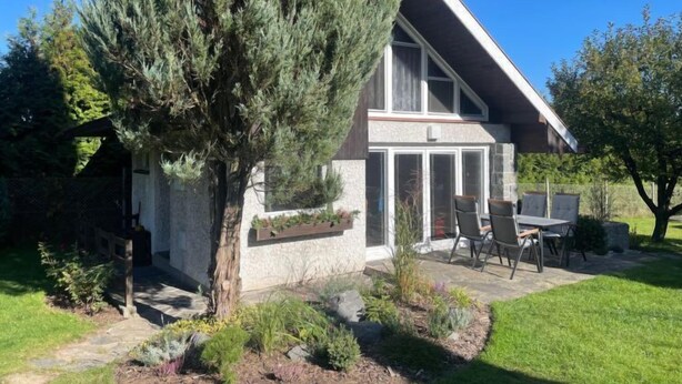Een modern huis met een stenen gevel, een grote boom in de voortuin, en een terras met tuinmeubilair.