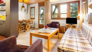 A cozy living room with wooden furniture, a dining area, and large windows offering a view of the outdoors.