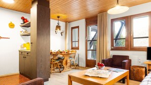 A cozy living room with a wooden table, chairs, and a sofa. There is a dining area with a wooden table and chairs. The room has wooden floors and walls, and a large window with curtains.