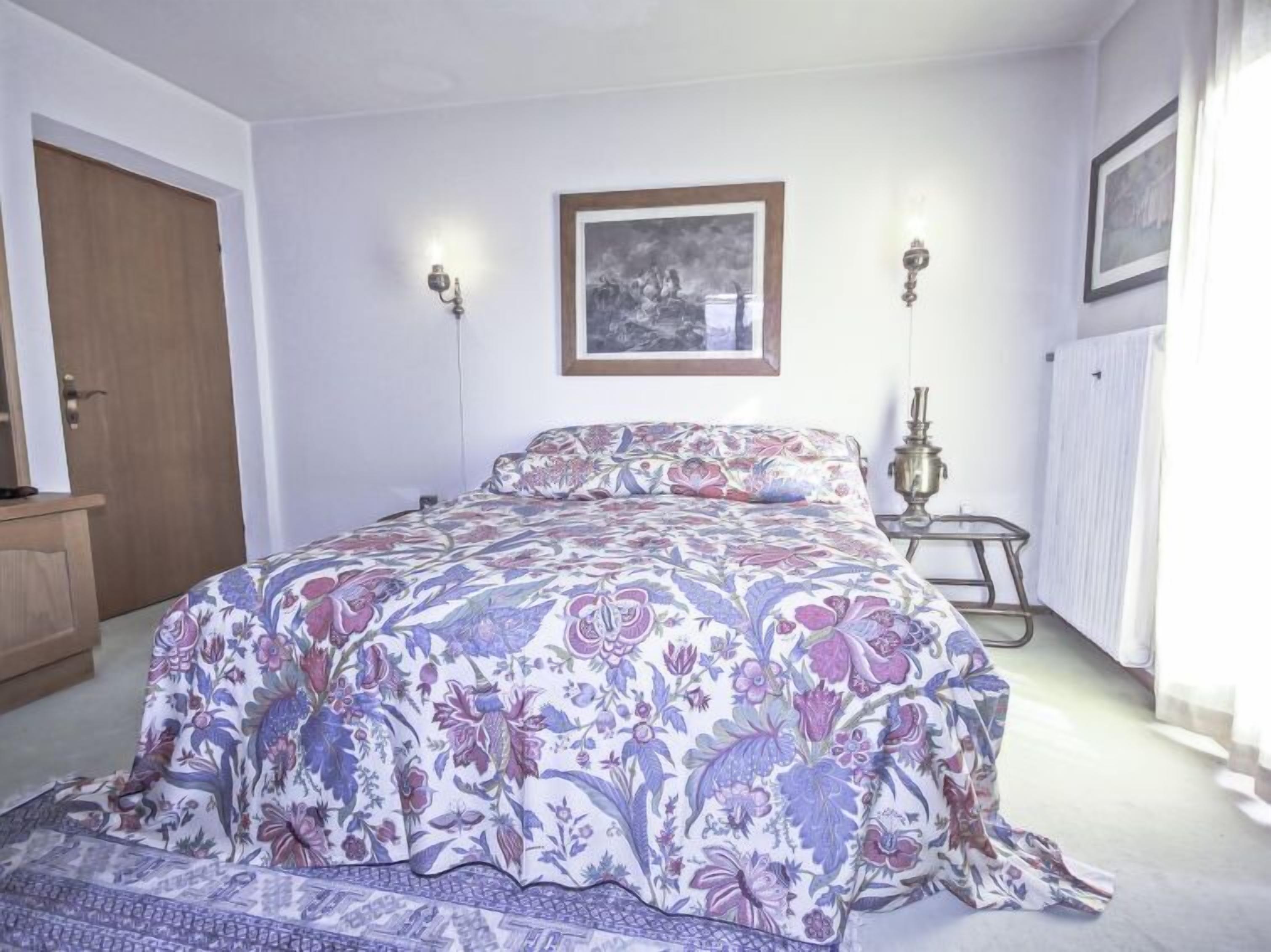 Une chambre à coucher avec une couvre-lit à motifs floraux, une porte en bois, un tableau encadré au mur et une petite table avec une lampe.
