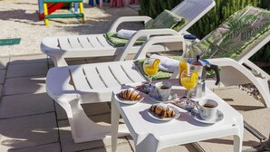 Un espace de détente aménagé sur la plage, avec des chaises longues blanches, une table où sont servis des articles pour le petit-déjeuner, notamment des croissants, du café et du jus d’orange, ainsi qu’une bouteille de jus.